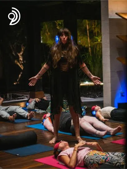 Woman leading a silent yoga session with participants on yoga mats wearing headphones.